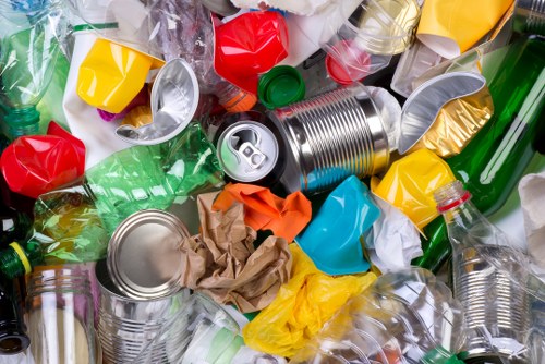 A cluttered pile of mixed recyclable waste, predominantly consisting of crumpled paper, crushed aluminium cans with metallic finishes and open mouths, and transparent plastic bottles with varying cap colours including red, green, and clear. The waste is scattered across a paved surface, likely an outdoor area or driveway, with some containers slightly deformed from compression. The background shows additional plastic bottles and packaging, indicating a collection of refuse awaiting rubbish removal services. The overall scene highlights a range of packaging materials, including plastic and aluminium, in different textures and colours, associated with waste collection activities in Finchley or surrounding areas. The presence of the waste materials demonstrates the type of refuse that Commercial Waste Finchley might handle as part of their rubbish removal services.
