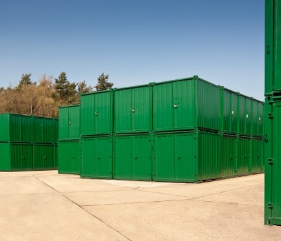 A large area of stacked green metal shipping containers arranged in a rectangular formation on a concrete surface, with some containers placed directly on the ground and others elevated slightly, set against a backdrop of trees and a clear blue sky. The containers have a ribbed texture with visible handles and locking mechanisms on the doors. The scene appears to be part of a commercial waste storage or disposal yard, likely in Finchley or nearby London, as part of a rubbish removal service by Commercial Waste Finchley. The containers' vibrant green colour and the orderly stacking suggest a professional setup for waste management, with no surrounding vehicles or additional equipment visibly present in the foreground. The environment is well-lit with natural daylight, indicating a clear day, suitable for waste logistics operations typical in the Finchley area.