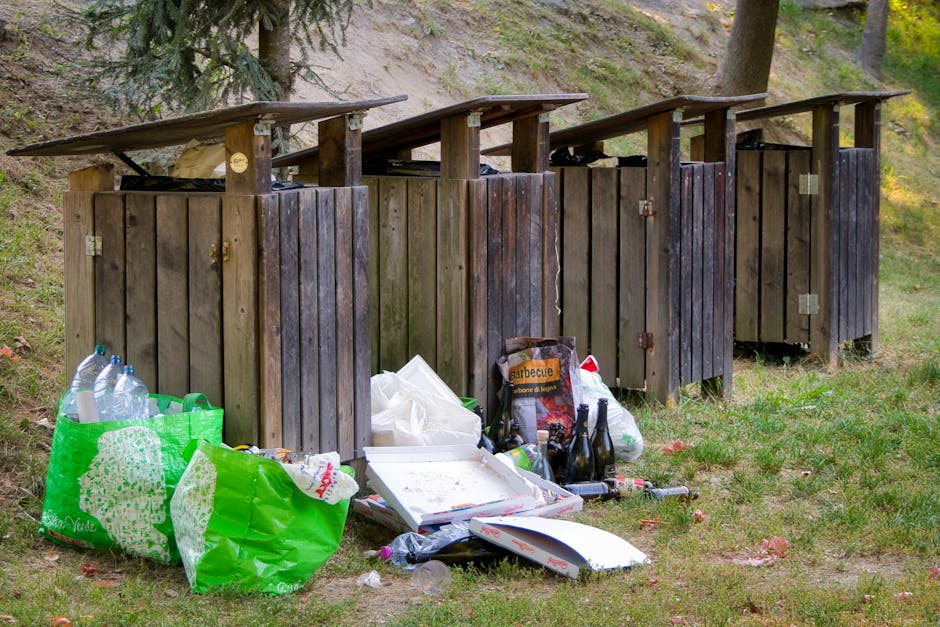 A row of four wooden outdoor rubbish bins with sloped roofs, positioned on a grassy area near a sloped, tree-lined hillside. The bins are made of weathered, dark brown and lighter wood planks, with me
