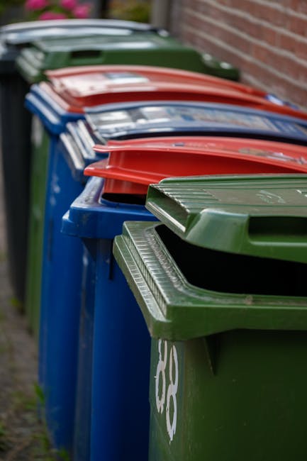 A row of four wooden outdoor rubbish bins with sloped roofs, positioned on a grassy area near a sloped, tree-lined hillside. The bins are made of weathered, dark brown and lighter wood planks, with me