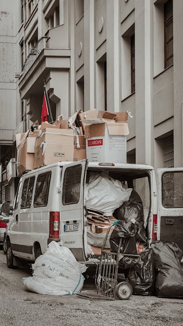 A burgundy flatbed utility vehicle parked on a city street in front of a modern office building with large windows and concrete paneling. The truck's cargo area is filled with various waste materials,